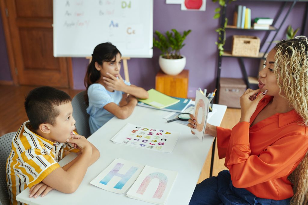 Teacher engaging with young students in classroom setting teaching letters. Child attentively focused on learning material with colorful visuals displayed on table